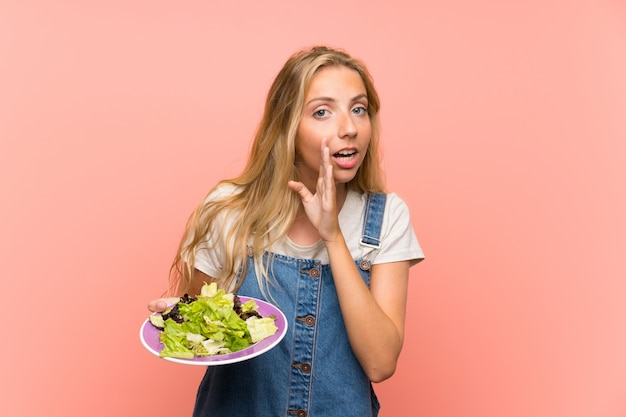 Blonde young woman with salad over isolated pink wall whispering something