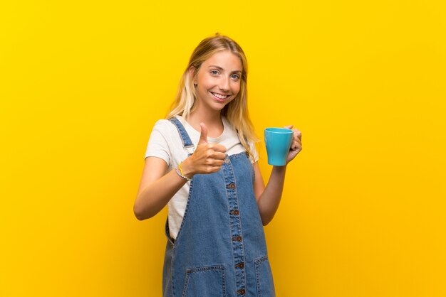 Blonde young woman over yellow wall holding hot cup of coffee