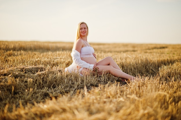 Blonde zwangere vrouw op kroongebied bij witte ondergoedkleren op zonsondergang