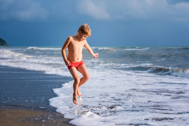 Blondejongen die en op het strand op blauwe overzeese kust in de zomervakantie in de dagtijd lopen springen.