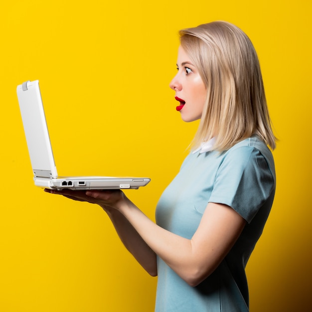 Blondemeisje in blauwe kleding met laptop computer op gele ruimte