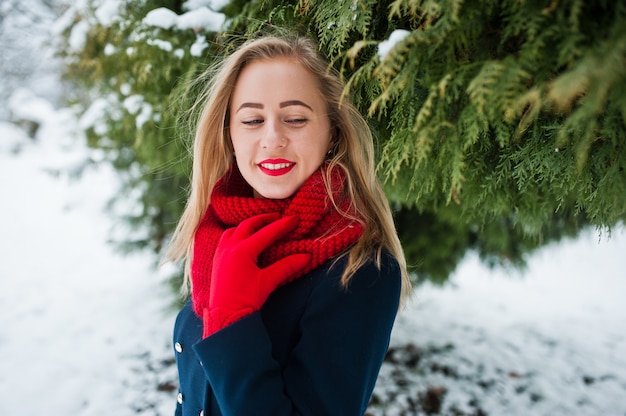 Blondemeisje in rode sjaal en laag die bij park op de winterdag lopen.
