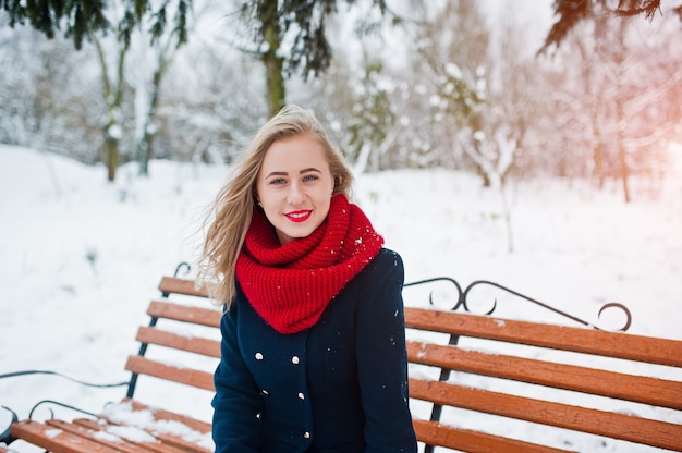 Blondemeisje in rode sjaal en laagzitting bij bank op de winterdag.