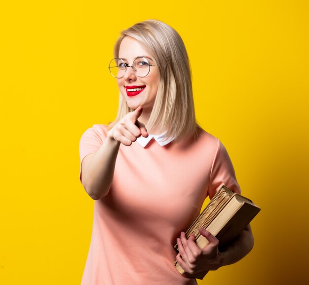 Blondemeisje in roze kleding en glazen met boeken op gele ruimte