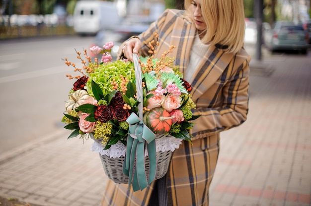 Blondevrouw die een rieten mand van de herfstbloemen houden tegen de stad