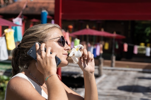 Blondevrouw met zonnebril die een witte lelie ruiken terwijl het spreken op haar mobiel voor een gebouw