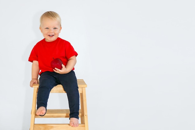 A blondhaired child in a red Tshirt smiles holding an apple in his hand sitting on a chair