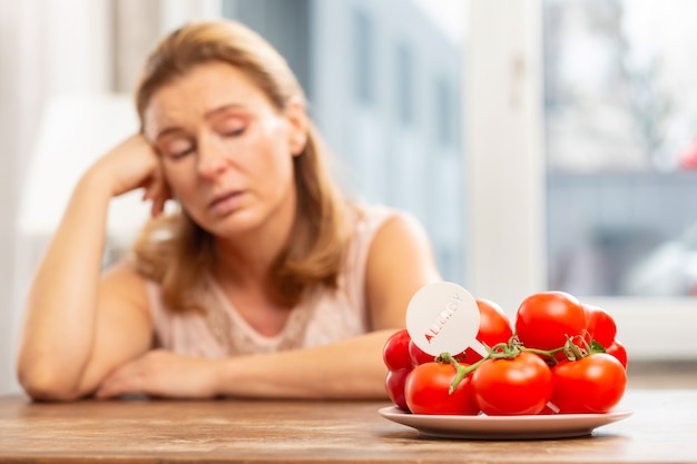 Blondharige huisvrouw zit aan tafel en kijkt naar tomaten maar heeft een allergie