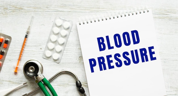 BLOOD PRESSURE is written in a notebook on a white table next to pills and a stethoscope