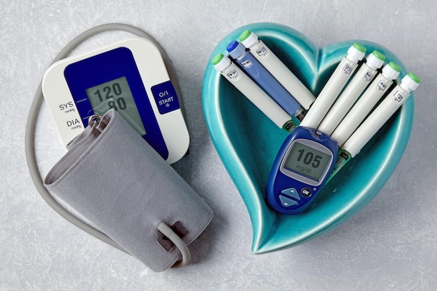 Blood pressure monitor blood glucose monitor insulin syringe\
pens in a blue heartshaped bowl on a light gray concrete table\
medical equipment for health monitoring and therapy