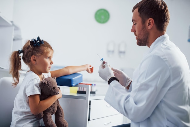 Photo blood sampling little girl with her teddy bear is in the clinic with doctor