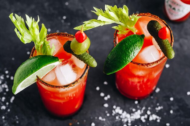 Bloody Mary Cocktail in glasses with garnishes Tomato Bloody Mary ice cold drink with fresh celery pickles and lime on dark background copy space