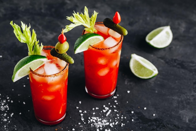 Bloody Mary Cocktail in glasses with garnishes Tomato Bloody Mary ice cold drink with fresh celery pickles and lime on dark background copy space