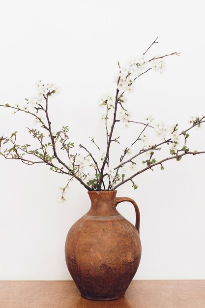 白い壁に木製のテーブルに咲く桜の枝 ビンテージ花瓶の静物画の春の花 シンプルな田舎暮らしの家の素朴な装飾 テキスト用のスペース