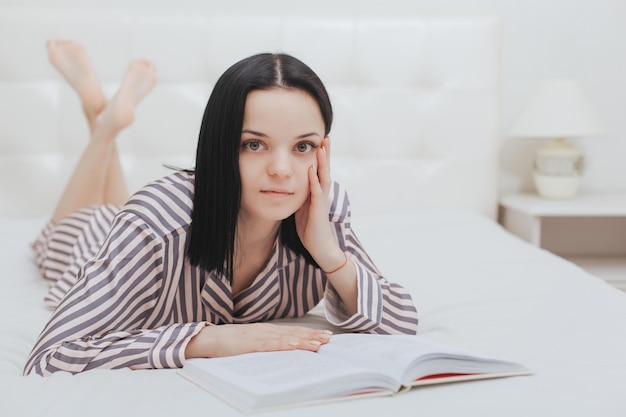 Blootsvoets jong tienermeisje dat naar de camera kijkt die op haar bed ligt en een boek leest