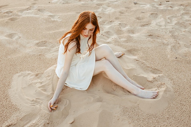 blootsvoetse vrouw met golvend rood haar en in jurk zittend op zandstrand