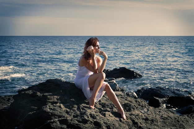 Blootvoetse dame die naar shell luistert terwijl ze op een rotsblok aan zee zit