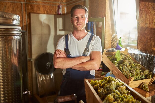 Foto blozende jonge man met zijn armen gekruist terwijl hij in de buurt van containers met druiven bij de wijnmakerij staat