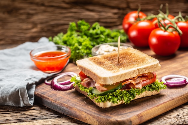 Photo blt sandwich on a cutting board next to sweet chilli sauce and fresh vegetables.