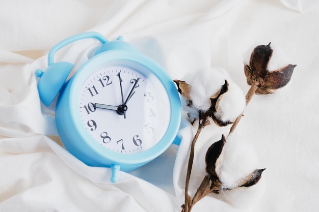 Синий будильник и цветок хлопка на кровати фоне белой мятой ткани