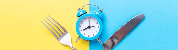 青い目覚まし時計、フォーク、色紙の背景にナイフ。断続的断食の概念。水平バナー-画像