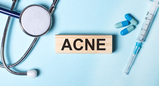 On a blue background, a stethoscope, a syringe and pills and a wooden block with the word ACNE. Medical concept