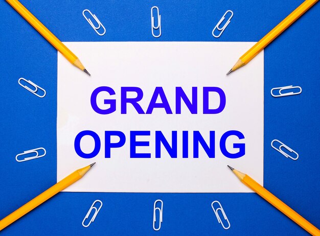 On a blue background, white paper clips, yellow pencils and a white sheet of paper with the text GRAND OPENING