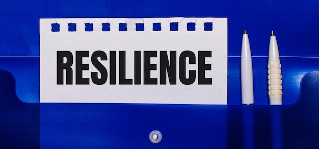 На синем фоне белые ручки и лист бумаги с надписью RESILIENCE.