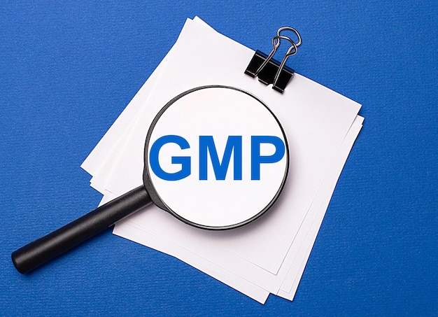 Photo on a blue background white sheets under a black paper clip and on them a magnifying glass with the text gmp good manufacturing practice