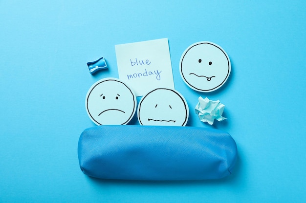 Синяя сумка и бумаги с грустными смайликами и текстом Blue Monday на синем фоне, вид сверху