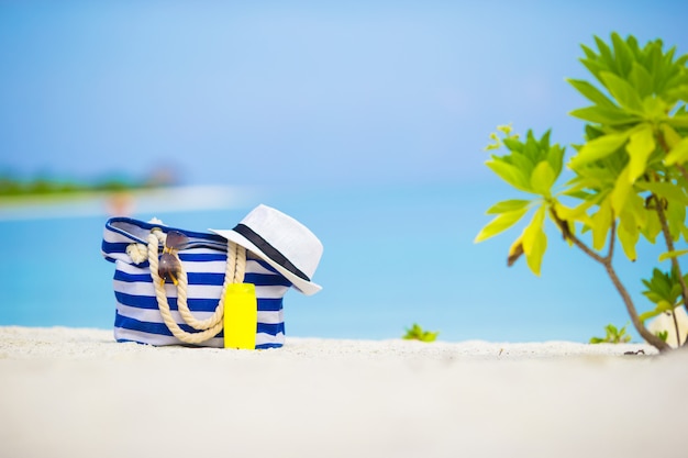Borsa blu, cappello di paglia, occhiali da sole e bottiglia di crema solare sulla spiaggia bianca