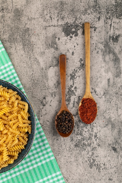 Blue bowl of dry raw fusilli pasta and various condiments on stone table. 