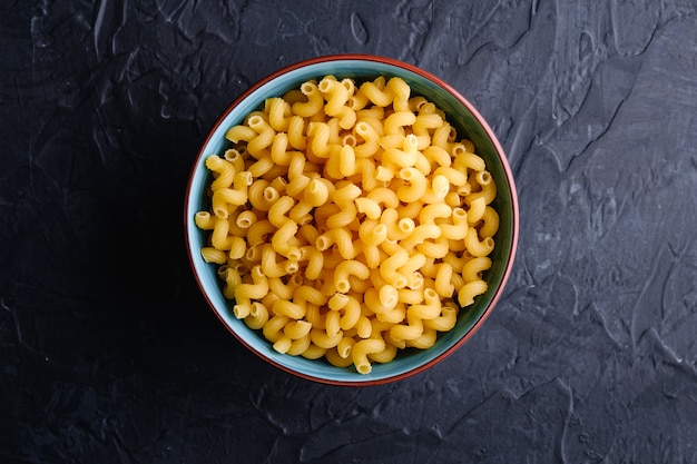 Blue bowl with cavatappi uncooked golden wheat curly pasta on textured dark black table, top view