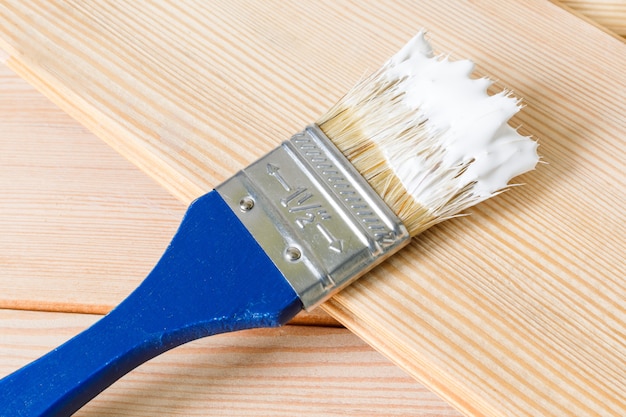 Синяя кисть в белой краске лежит на легкой деревянной доске. Начало ремонтных работ в доме. Выборочный фокус, копирование пространства