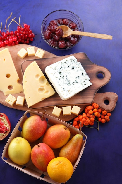 Blue cheese and yellow hard cheese with fruits and berries on wooden boards Various types of cheese composition with honey and apples in a wooden basket on a blue background