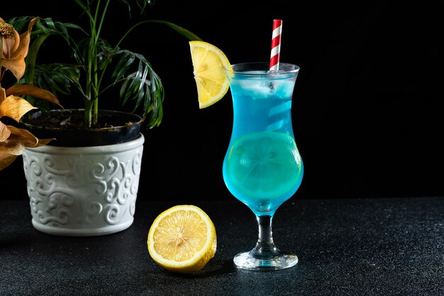 A blue cocktail glass decorated with Sicilian lemon, vase with a small palm tree next to the drink, on a dark background