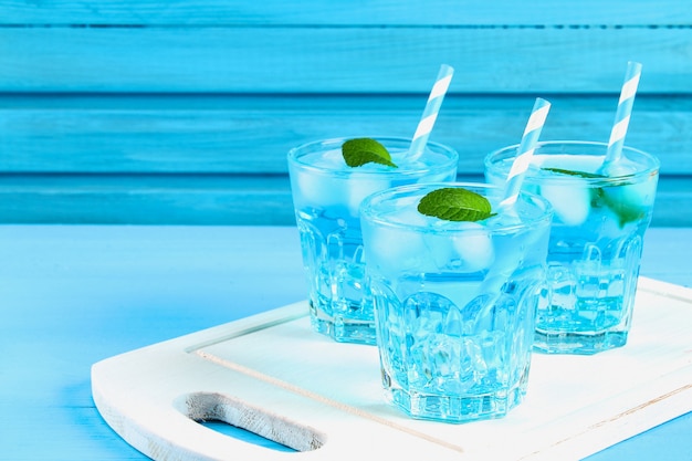 Blue cocktail with ice and mint in glasses on a white wooden board on a blue table.