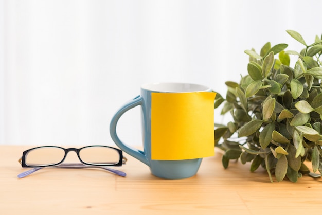 Blue coffee cup, eyeglassses and yellow post it for text on wood table top