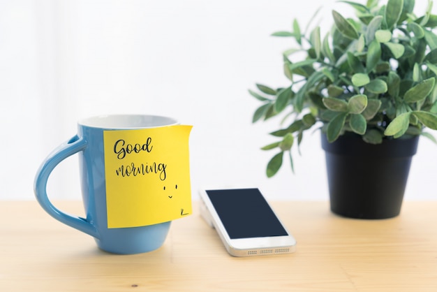 Blue coffee cup and post-it note with message "Good Morning", on wooden table