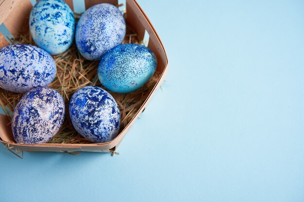 Blue cosmos chicken eggs lie in round wooden basket which stands on a blue background.