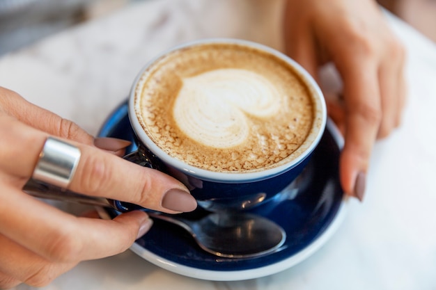 Blue cup with cappuccino with heart-shaped foam in female hands at a table in a cafe. Close-up. Soft focus.