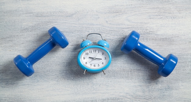 Blue dumbbells and alarm clock on white wooden background.