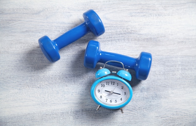 Blue dumbbells and alarm clock on white wooden background.