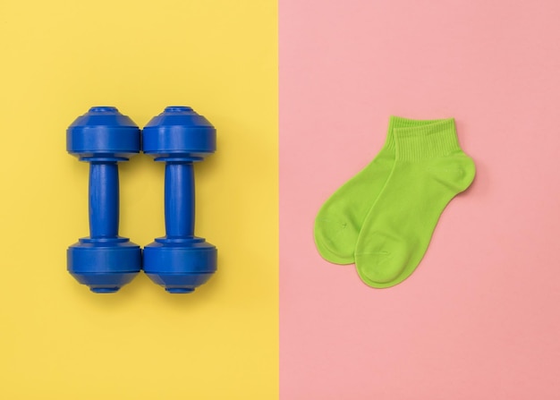 Blue dumbbells and green socks on a pink and yellow background. Accessories for fitness and weightlifting.