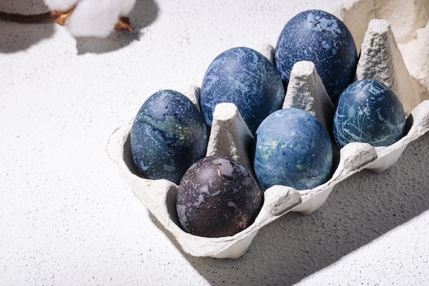 Blue easter eggs dyed with hibiscus tea in an egg carton on a\
white concrete background