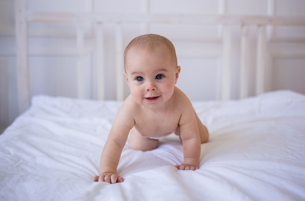 Blue-eyed baby girl in a diaper crawls on a white cotton blanket on the bed in the room