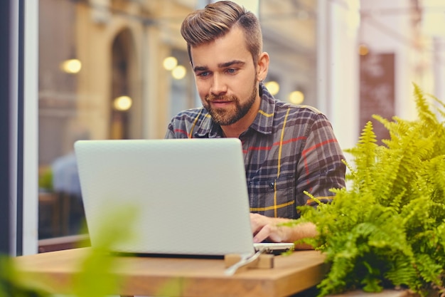 Foto maschio barbuto e dagli occhi azzurri che usa un laptop in un caffè in una strada.