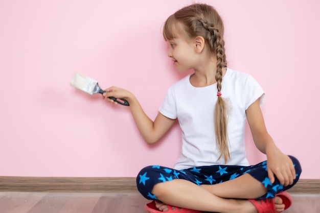 白いTシャツを着た青い目のブロンドの女の子が笑顔でピンクの背景に子供部屋の改修のためのブラシを保持しています