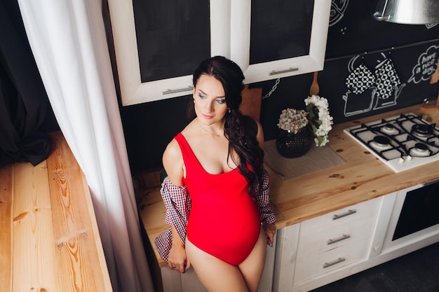 Blue eyed brunette woman in red swimming suit shirt and sneakers leaning on furniture Young pregnant lady with dark long hair standing in kitchen Happy stylish model smiling and posing at camera