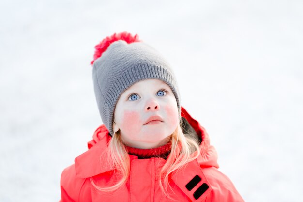 A blue-eyed little girl in a knitted hat and a pink winter jacket looks up. 